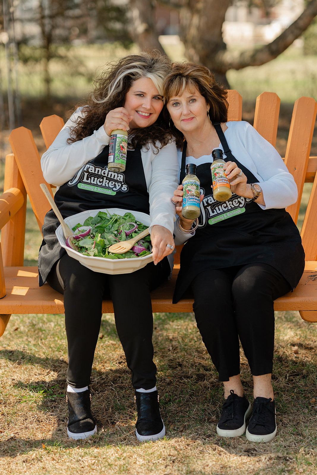 Two women (the owners - mother and daughter) sitting on a bench outdoors, holding bottles and a salad.