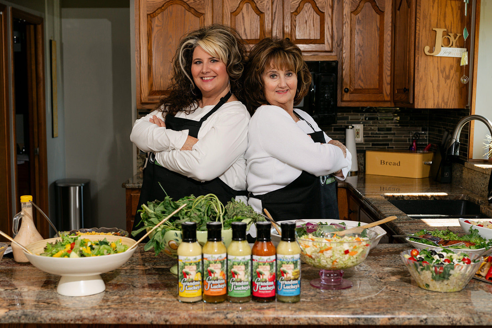 Mother and daughter owners in a kitchen with salad ingredients and dressings on a counter.