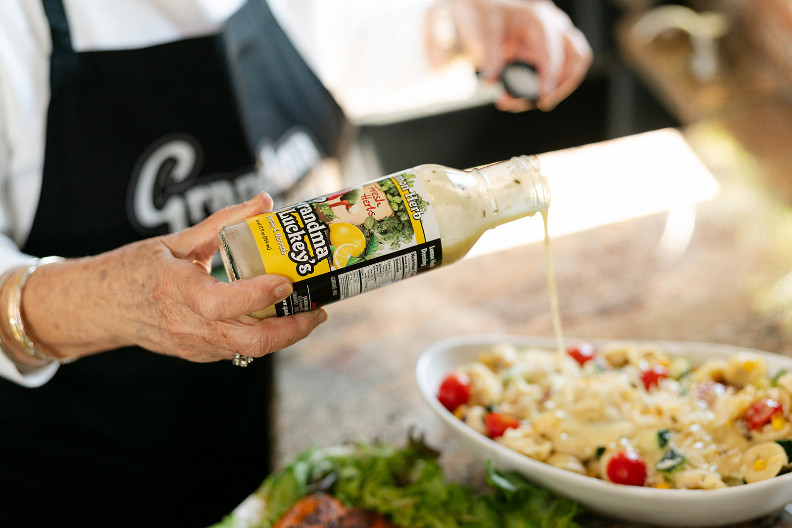 Women pouring salad dressing lemon herb from a bottle into a bowl of pasta salad.